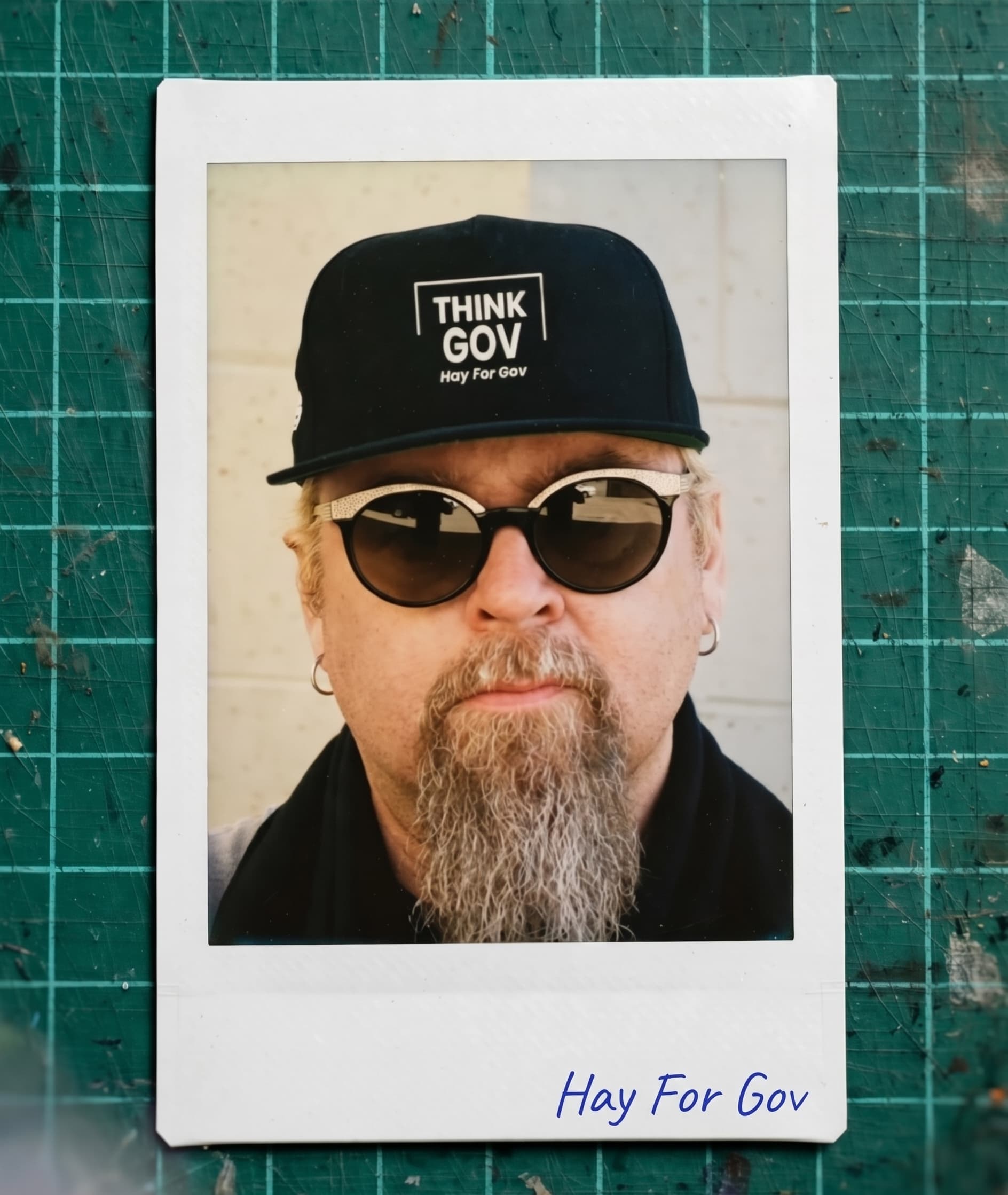 Polaroid of a man with a grey goatee, sunglasses, and a black "THINK GOV" cap.